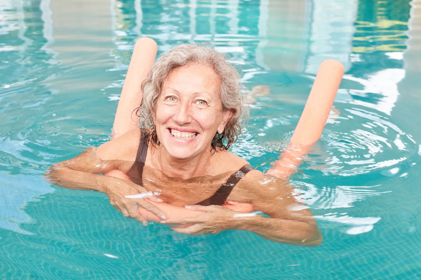 A smiling adult floating in a bright pool with orange pool noodles
